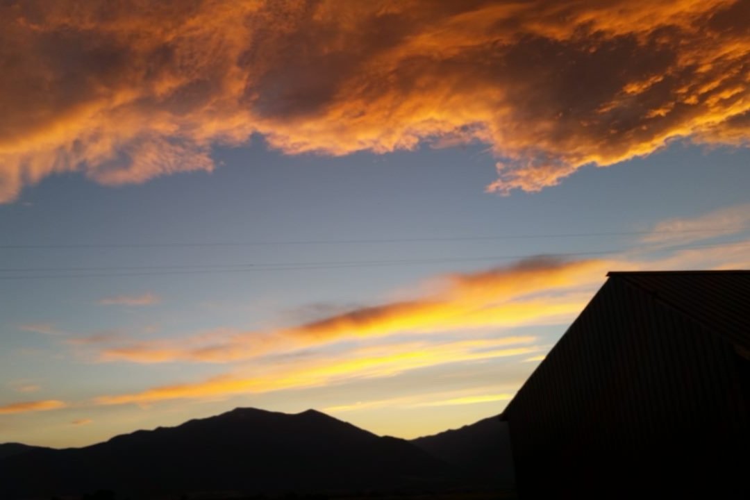 Beau+McLean+-+20180824_203603 Scenic sunset view with vibrant orange and yellow clouds over a mountain silhouette and a barn roof in the foreground.