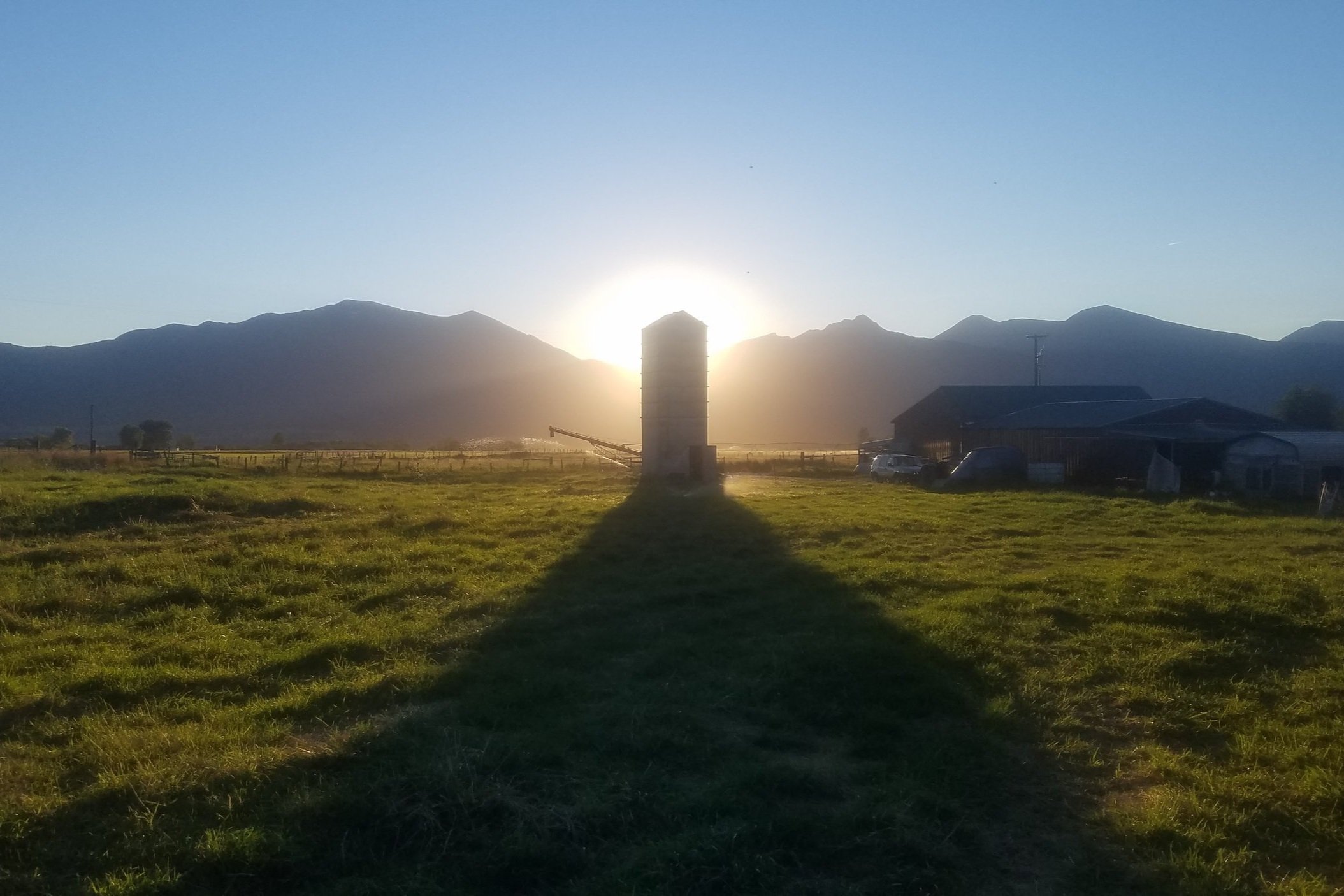 Beau+McLean+-+20200810_202406 Sunset view over a farm with a silo casting a long shadow, surrounded by green grass and mountains in the background, creating a serene rural landscape.