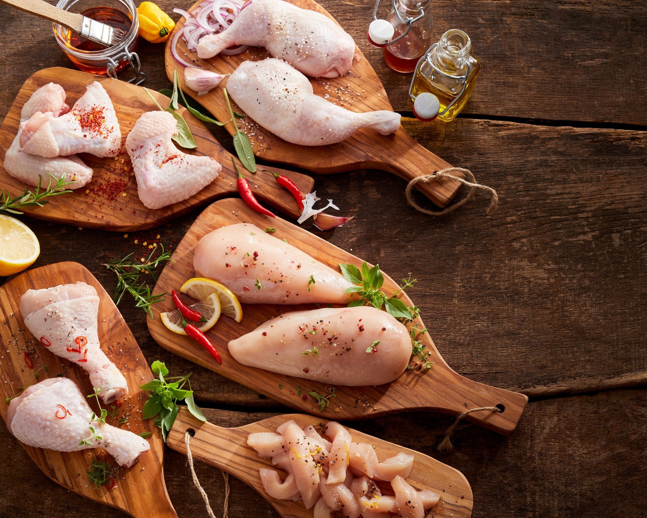 Assorted raw chicken cuts, including breasts, thighs, and drumsticks, arranged on wooden cutting boards with fresh herbs, spices, and lemon slices, set against a rustic wooden background.