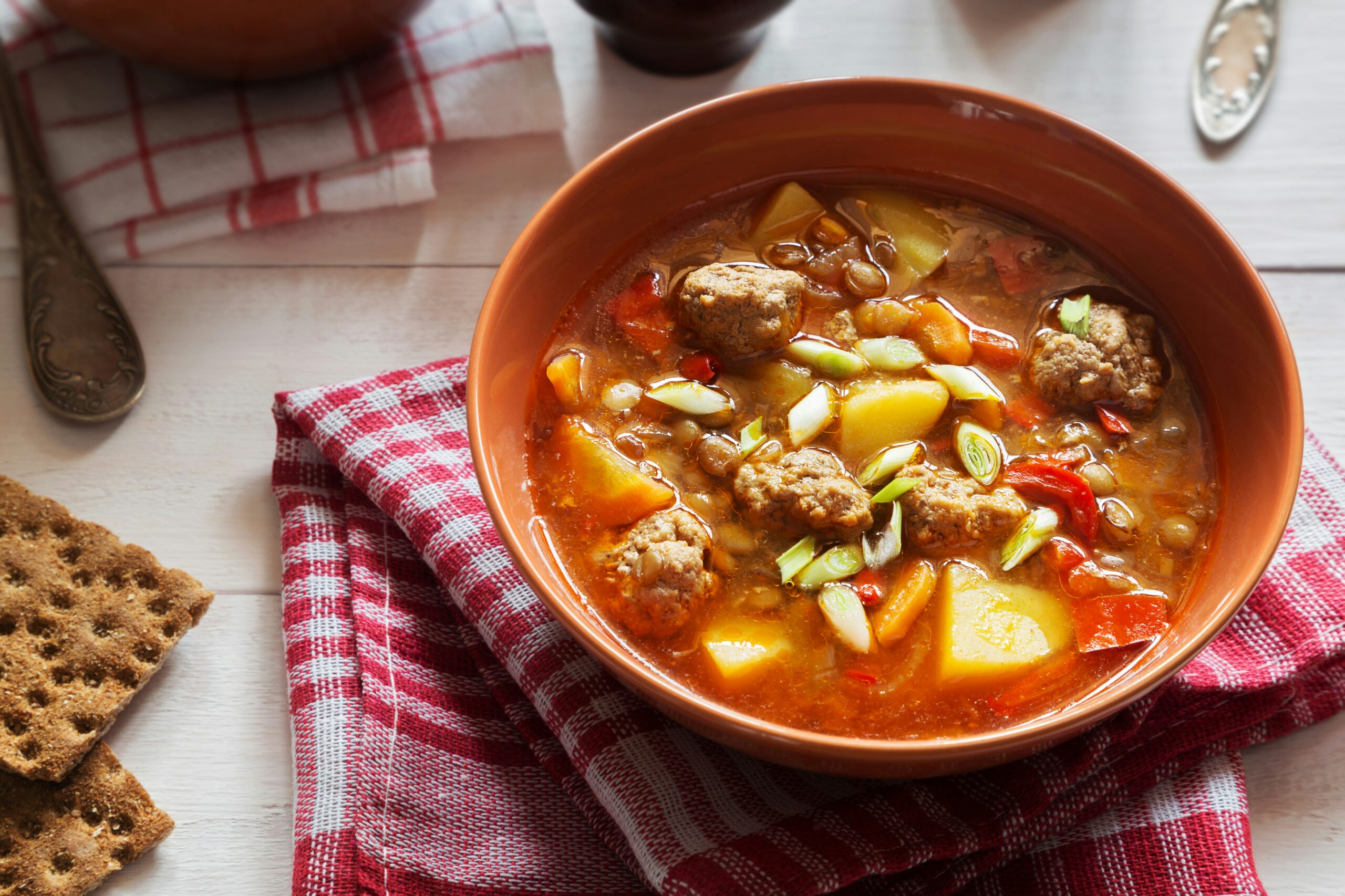 Bowl of hearty meatball soup with potatoes, vegetables, and green onions, served on a red and white checked napkin with whole grain crackers on the side.