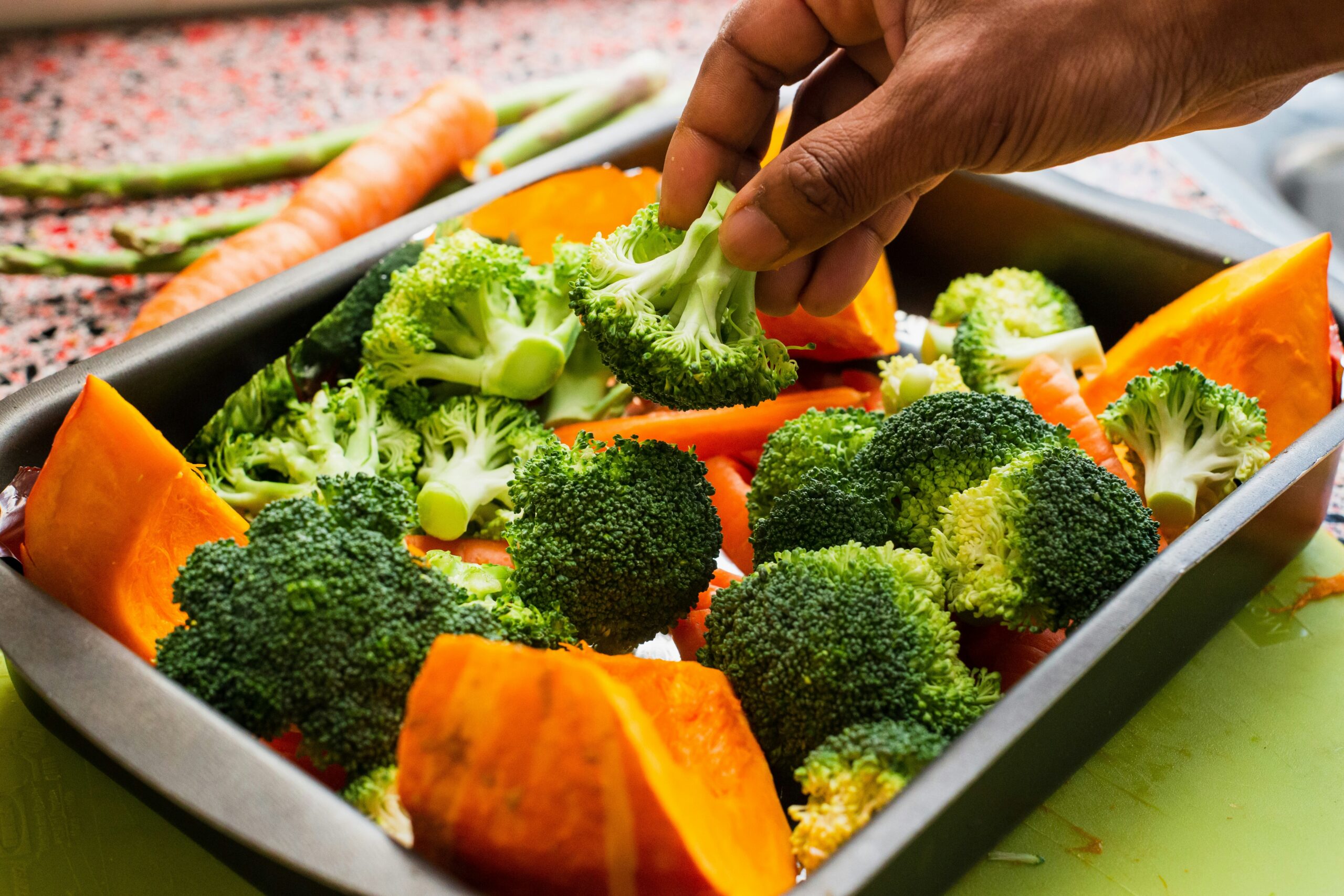 louis-hansel-TBKh5z8eKHY-unsplash Fresh broccoli, pumpkin, and carrots are arranged in a baking tray, with a hand selecting a piece of broccoli. The vibrant vegetables are ready for roasting, showcasing healthy meal preparation.